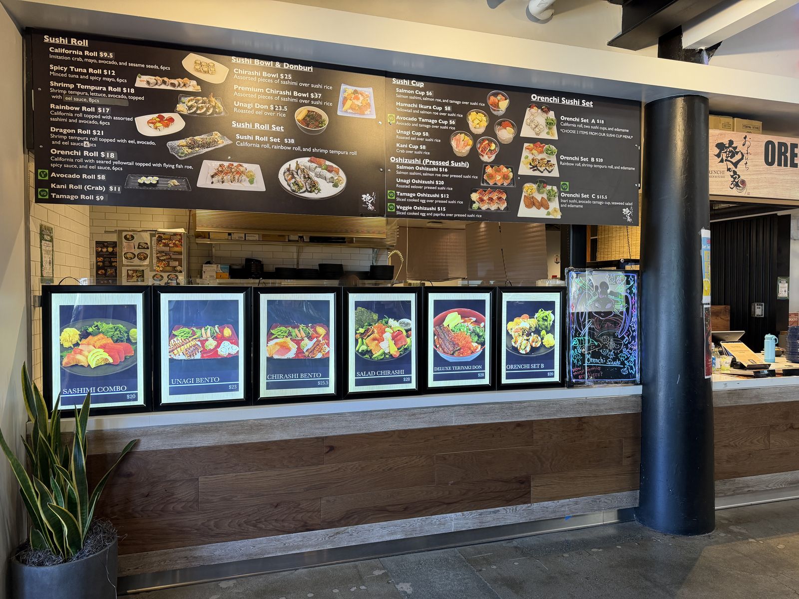 The Orenchi Sushi stall inside State Street Market, with an overhead sushi menu board and lit photo panels of sashimi combos and chirashi bowls along the counter